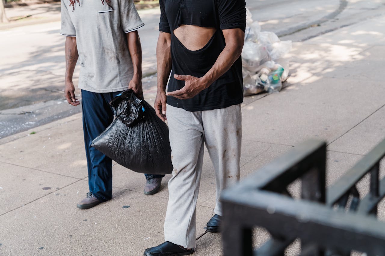 Two adults in worn clothes carrying a garbage bag, depicting poverty on a city street.